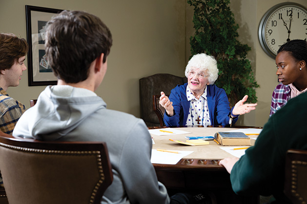 A senior citizen talks to three high schoolers at a table.