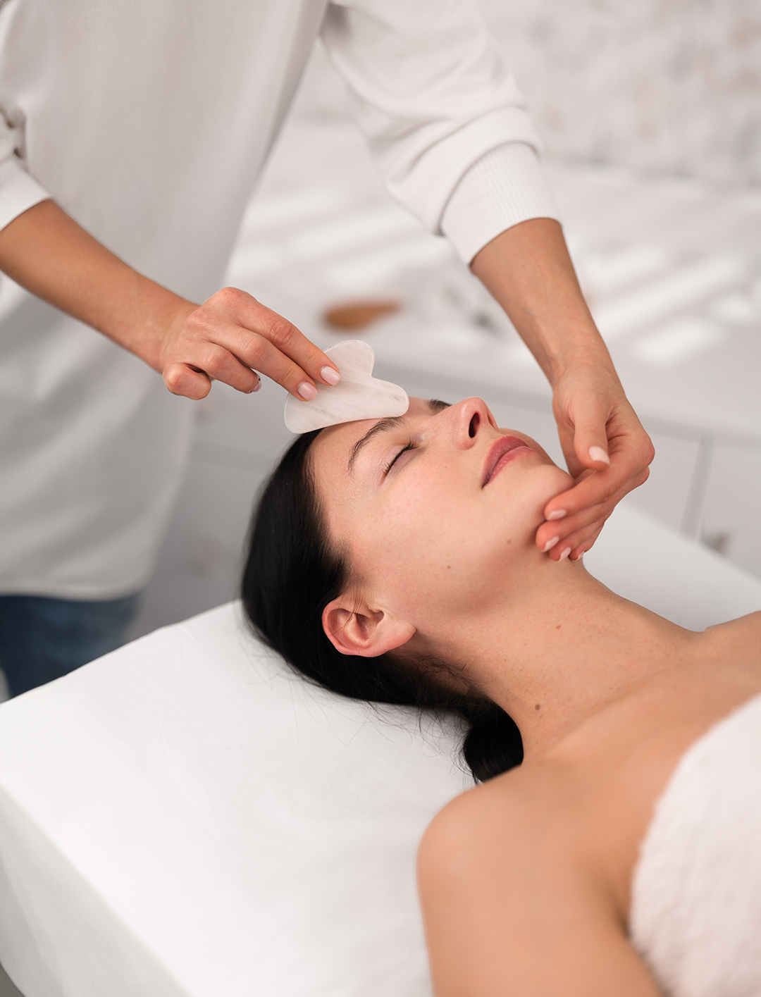 From above of crop unrecognizable cosmetician using gua sha tool while massaging face of peaceful young female client lying on couch with closed eyes